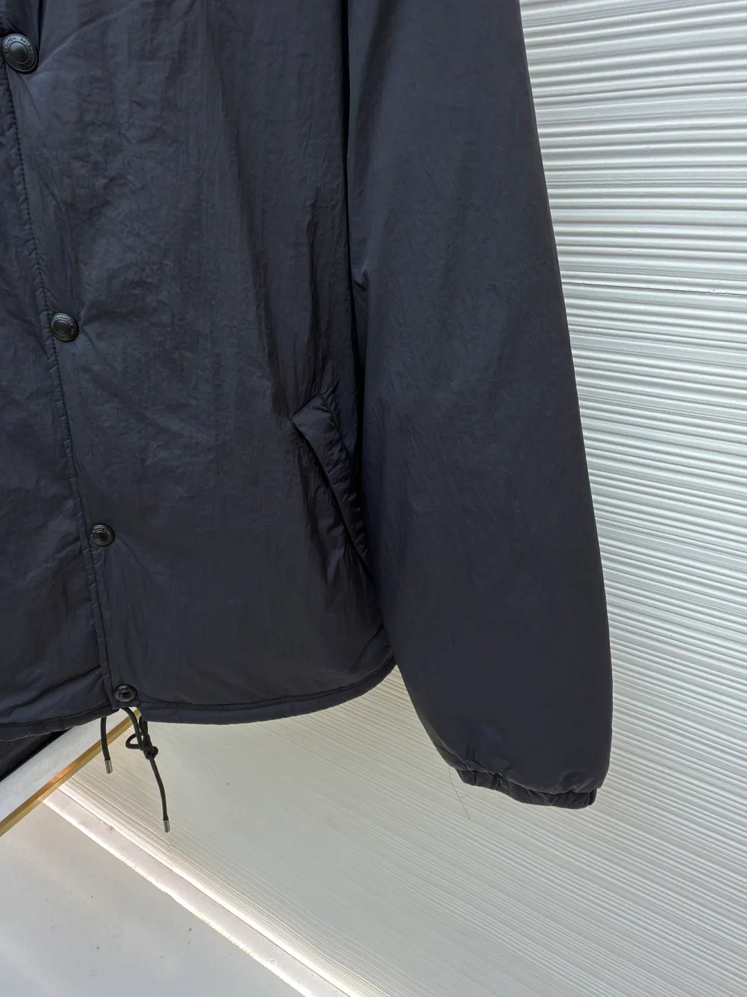 Ralph Lauren red label light cotton jacket (coach jacket) ●Original version: purchased and developed for 2699 ●Item number: 1681910-B90 ●Fabric: high-density nylon taffeta fabric ●Technology: embroidery production ●Color: black ●Version: fitted version ●Weight: about 340 grams ●Size: S-M-L-XL ●Description: The density of high-density nylon taffeta is about 210T-300T, with a smooth and slightly matte surface, excellent windproof and waterproof properties (with DWR water-repellent treatment), strong wrinkle resistance and easy care, in line with the positioning of American casual "durable and practical". Classic dark black, using vat dyeing process (dye penetrates into the fiber), the color fastness reaches ISO level 4 or above, preventing fading or color transfer after washing. The lightweight cotton-polyester blend lining (65% Polyester + 35% Cotton) reduces the friction between the fabric and inner clothing, improving the smoothness of wearing; the blended material combines breathability and durability to avoid the stuffy feeling of pure nylon lining. The classic Coach Jacket has a loose fit with slightly dropped shoulder lines (shoulder width is 1-2cm wider than the standard fit). The length reaches the hips and the sleeves are slightly tapered straight sleeves, taking into account both freedom of movement and casual style. Polo horse logo on the left chest: flat satin stitch embroidery (satin embroidery), embroidery thread: 100% polyester high color fastness embroidery thread (the color number is consistent with the brand's standard red color card), stitch density 12-15 stitches/cm, the outline of the horse logo is clear, and the details of the rider and horse are burr-free. Embroidery positioning: precise positioning through the template (12cm from the neckline and 8cm from the placket) to ensure left and right symmetry and centering. Topstitching technology: The seams of the body are made of double-needle flat seams (needle pitch 3cm/10 needles), the stitches are smooth and straight, the stitching color is dark black (the same color as the fabric), and the stitch width is 0.5cm, which enhances firmness while forming simple decorative lines. The splicing of the front placket and the body: the placket is made of double-layer folding + flat seam splicing, with a thickness of about 0.3cm to ensure that the buttons are not easily deformed when fixed; the edges of the placket are cut with a hot knife to avoid burrs. The sleeves are spliced ​​to the body: the armholes are spliced ​​with round-head keyhole machine seams + flat seams. After splicing, the armholes have a natural curvature and no sense of tightness during movement; the height of the sleeves is about 8-10cm, which is in line with the loose characteristics of casual jackets. Main button: black urea resin button, matte surface treatment, buttonholes are hand-made keyholes (6 strands of cotton thread, keyhole density 8 stitches/mm), and the edges of the eyelets are smooth and without burrs. Collar buttons: The first button on the collar is a hidden button (diameter 12mm), which prevents the collar from opening and improves the neatness of wearing.